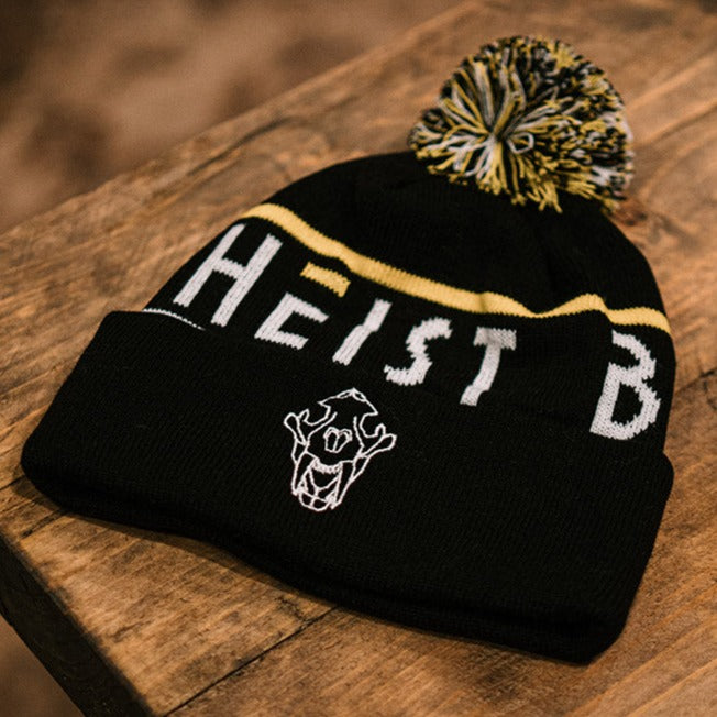 Black cotton bobble hat with white heist brew co logo and writing, yellow line around the top. White, black and yellow bobble on the top sat on a wooden table